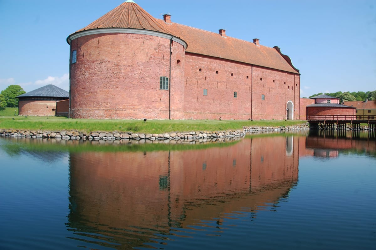 En stor, röd tegelbyggnad med ett runt torn står vid en vattensamling, med reflektioner av byggnaden synliga i vattnet. Gräsmattor och en stenskoning skiljer byggnaden från vattnet. En bro leder över vattnet till en mindre byggnad med rött tak.