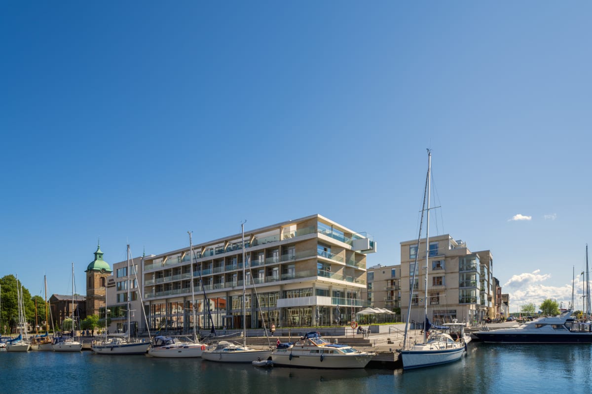 En bild som visar en marina med flera segelbåtar och en motorbåt förtöjda vid en kaj. Bakom kajen finns moderna byggnader med lägenheter eller hotell, och längre bort en äldre kyrkobyggnad med ett kopparbelagt torn. Himlen är klarblå med några små mo