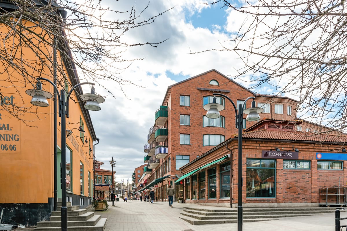 En gata inramad av nakna trädgrenar framför en rad byggnader i tegel och gult. Flera personer promenerar på gatan som kantas av gatlyktor.