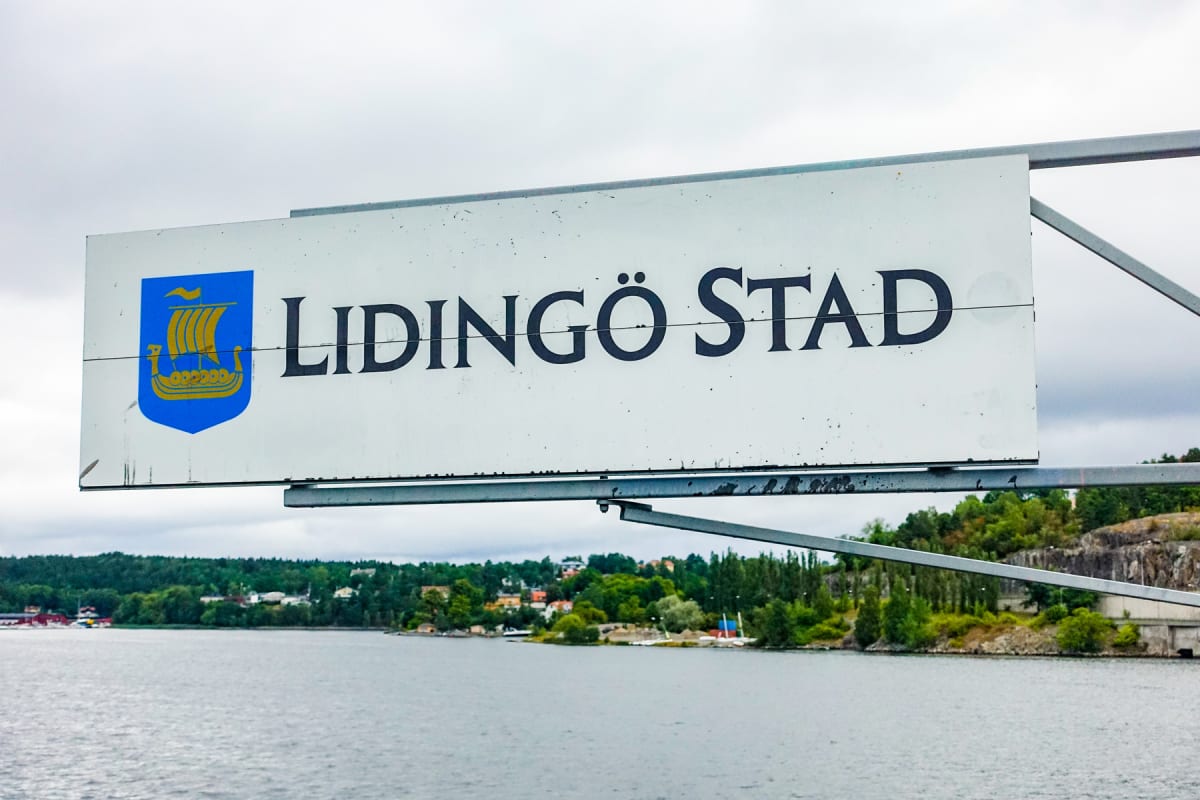 En skylt som avbildar Lidingö stads vapensköld med texten "LIDINGÖ STAD" syns i förgrunden. Bakom skylten ligger ett vattenområde med grönskande landskap och byggnader vid strandlinjen under en molnig himmel.