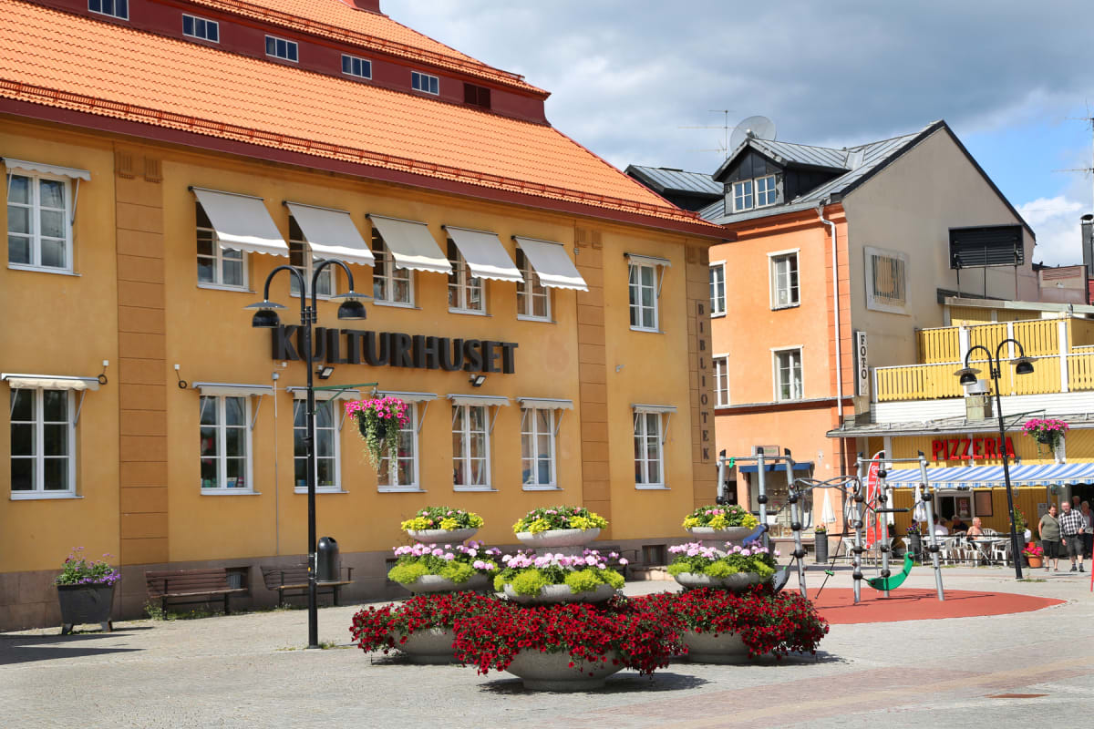 En bild som visar en central del av en stad med flera byggnader, inklusive ett "KULTURHUSET" och en pizzeria, flankerade av stora blomplanteringar. På höger sida syns en lekplats och uteservering. Byggnaderna har bland annat gula och orange fasader s