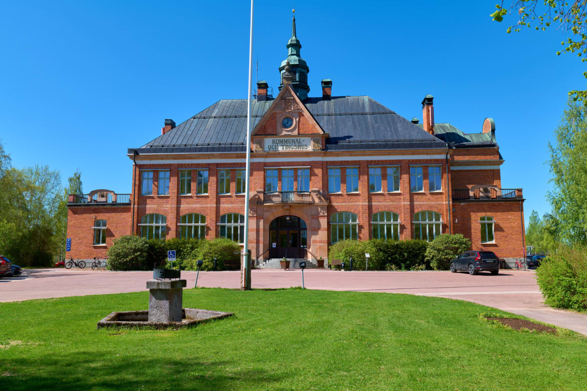 En stor, röd tegelbyggnad med ett mörkt tak och en spira i mitten. Framför byggnaden finns en gräsmatta, en flaggstång, ett stenfundament och en väg där en mörk bil och cyklar syns. Byggnaden har texten "KOMMUNAL- OCH TINGSHUS" på fasaden.