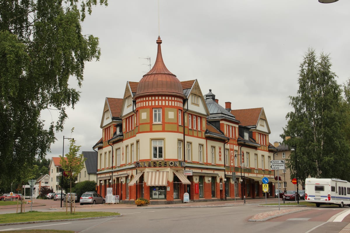 En stor, vacker herrgårdsliknande byggnad med en distinkt kopparkupol och röda tegeltak är central i bilden. En husbil står parkerad vid en vägkorsning, omgiven av träd och grönska, under en gråmulen himmel.