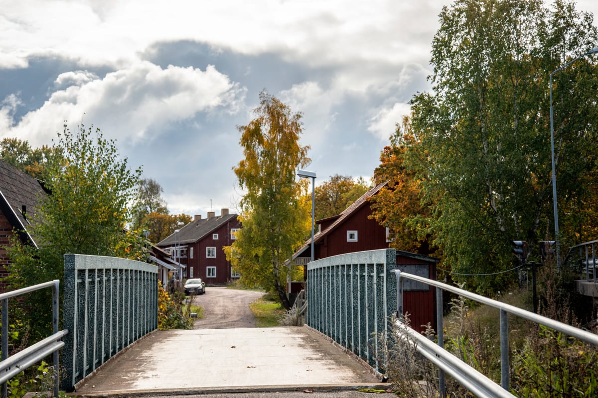 En bild tagen från en gångbro med metallräcken, som leder mot en grusväg. Runt vägen står flera röda trähus med vita fönster och mörka tak, omgivna av träd med löv i gröna, gula och orangea höstfärger. Himlen är täckt av moln.