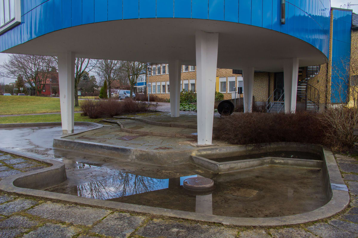 En utomhusbild som visar en modern byggnad med blå fasad och ljusgrå pelare som bär upp ett utskjutande takparti. Framför byggnaden finns en betongbassäng med reflekterande vattenpölar och en stensatt yta. I bakgrunden syns andra byggnader, träd och