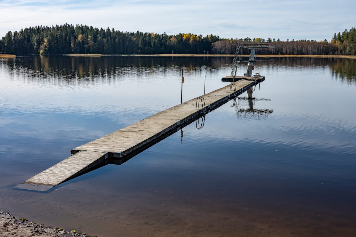En lång brygga med en badplattform och ett hopptorn sträcker sig ut över en lugn sjö under en delvis molnig hösthimmel. Träd med höstfärger syns på sjöns motsatta strandlinje.