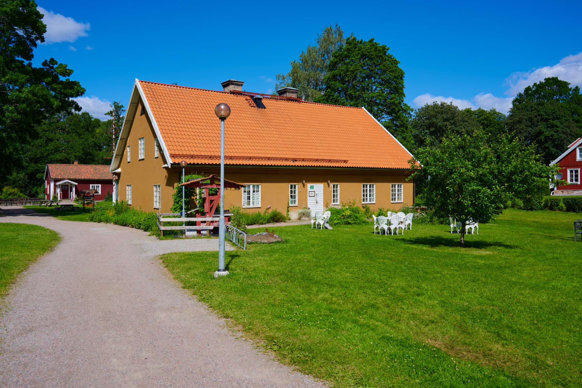 En gulmålad byggnad med tegeltak och vita fönsterkarmar står mitt i bilden. Framför byggnaden finns ett grönområde med vita utemöbler och en grusgång. I bakgrunden syns träd och delar av en röd byggnad.