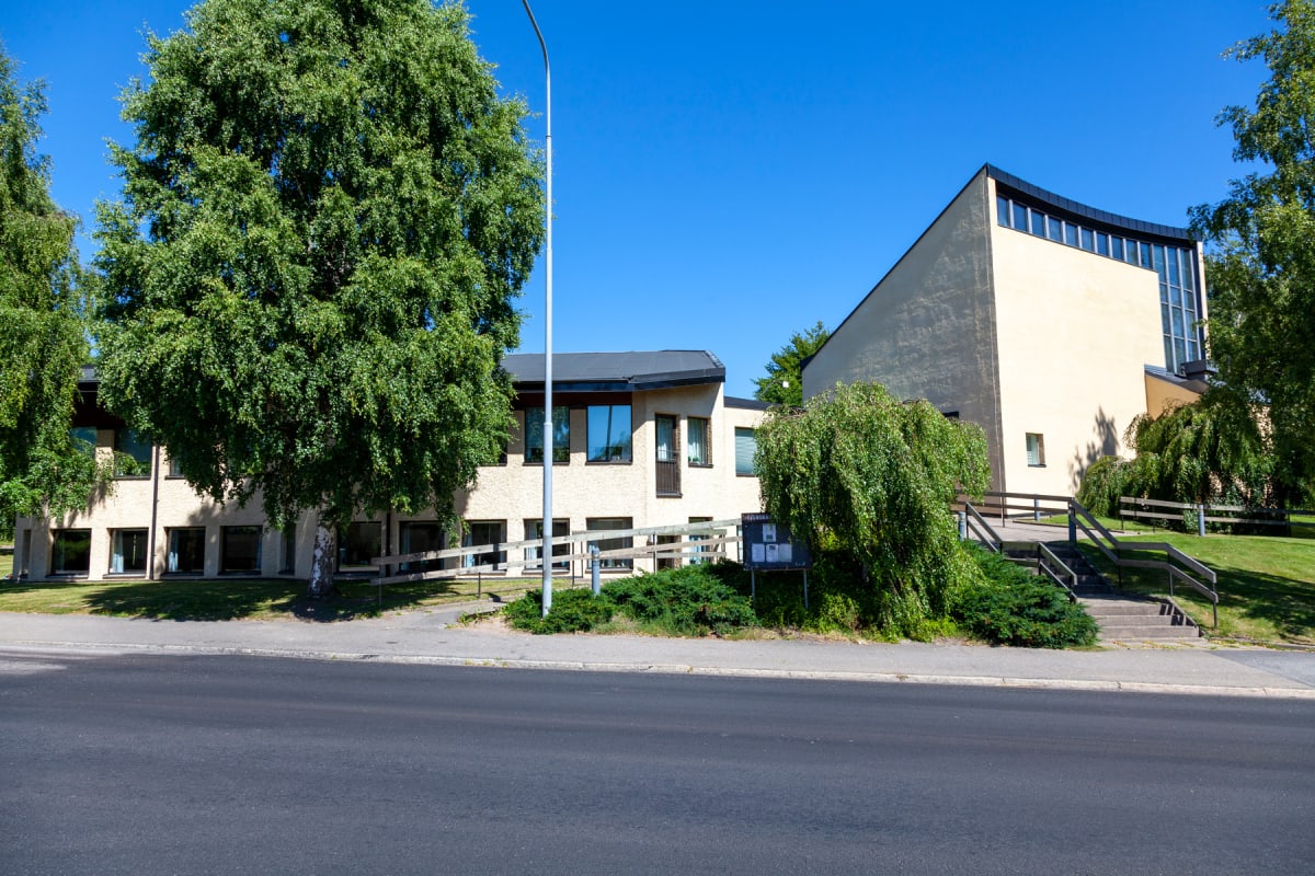 En modern byggnad med ljusgul fasad och mörkt tak, omgiven av grönska och träd under en klarblå himmel. Framför byggnaden finns en asfalterad väg och en gångväg med en anslagstavla och trappor som leder upp till ingången.