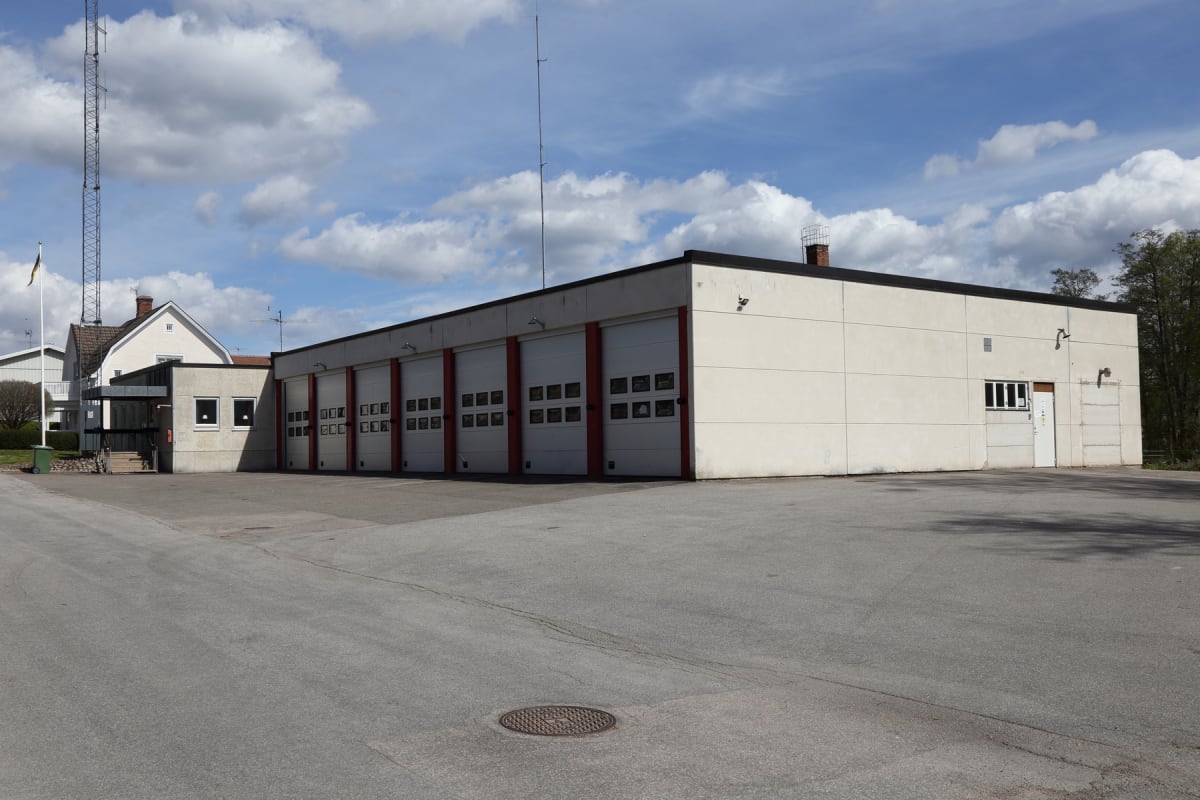 En modern byggnad med sex garageportar och en sidodörr är synlig under en delvis molnig himmel. Byggnaden har en ljus fasad och röda detaljer runt garageportarna. Framför byggnaden finns en asfalterad yta, i bakgrunden skymtar en radiomast och ett bo