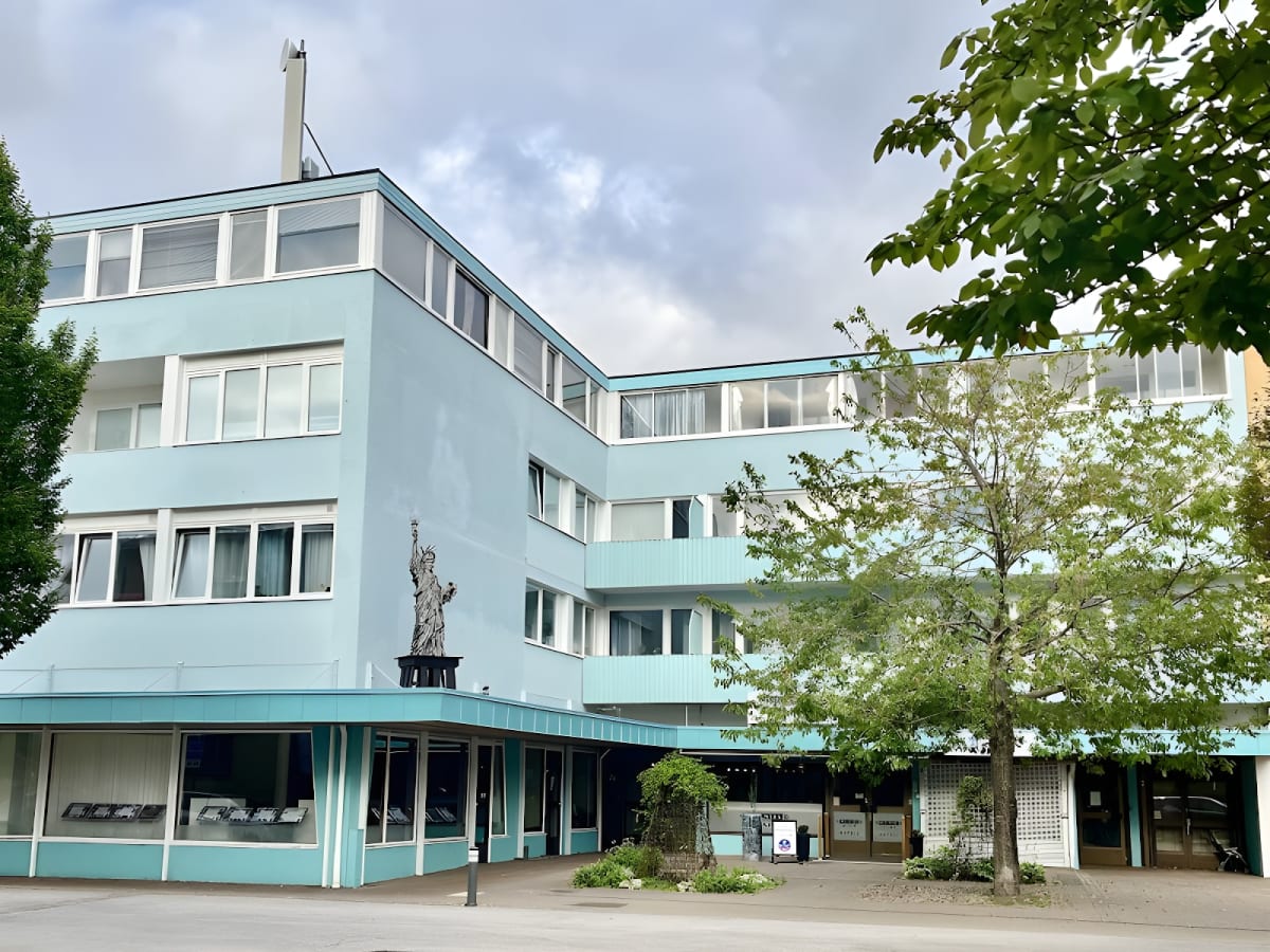 En ljusblå byggnad med flera våningar och många fönster syns under en molnig himmel. Framför byggnaden finns träd med gröna löv och på en avsats syns en staty föreställande Frihetsgudinnan. Markplan har stora skyltfönster.