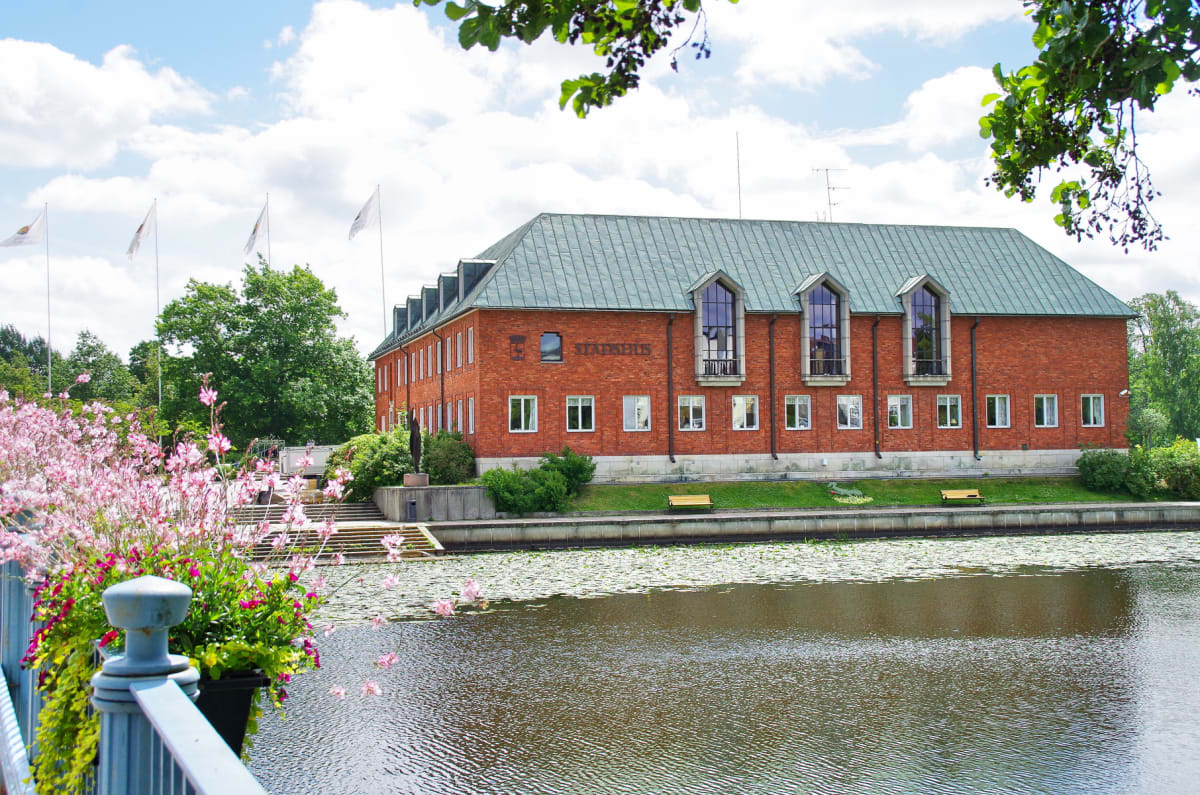 En röd tegelbyggnad med grönt tak och texten "STADSHUS" på fasaden, belägen vid en vattenyta med näckrosor. I förgrunden syns rosa blommor och ett blått räcke. På himlen syns vita moln och lövverk på träd.