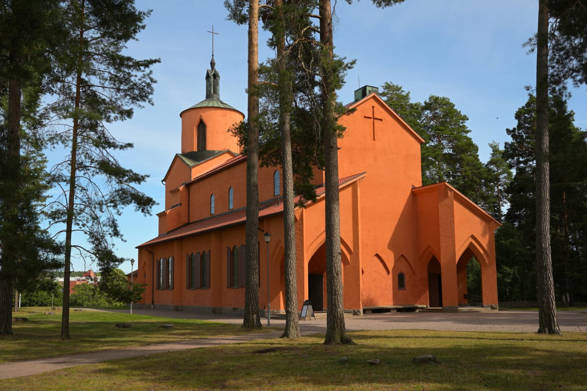 En stor, orange kyrkobyggnad med ett koppartak syns under en blå himmel, omgiven av gräsmattor och höga tallar. Kyrkan har en spetsig klocktorn med ett kors och flera valvformade fönster och dörröppningar. En grusgång leder förbi byggnaden.