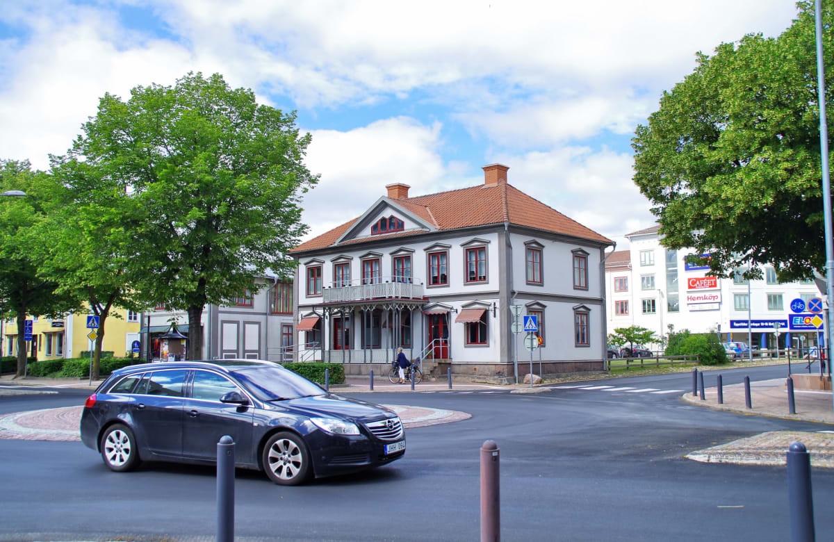 En svart stationära bil kör genom en rondell. På höger sida syns en stor, vit byggnad med rött tak och röda fönsterkarmar. I bakgrunden syns flera träd med gröna löv och byggnader under en delvis klarblå himmel med vita moln.