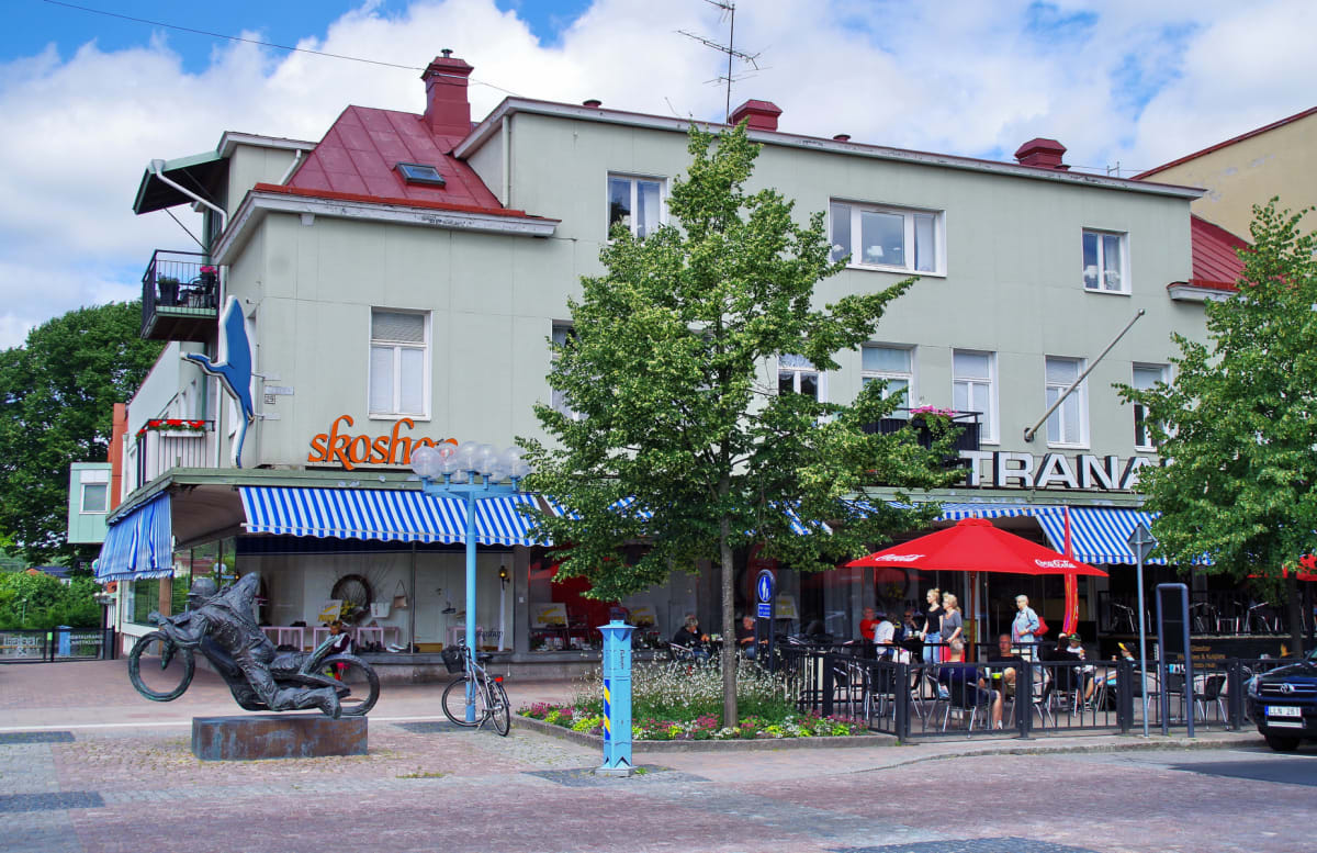 En bild visar en gata i en stad med en flervåningsbyggnad som inhyser affärer och restauranger på markplan. Framför byggnaden finns en terrass med bord och stolar under en röd parasoll, samt en staty föreställande en motorcyklist.
