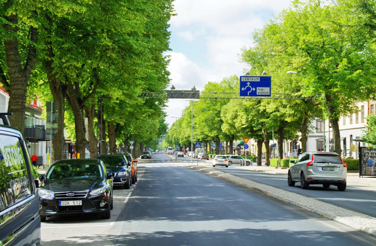 En gata kantad av gröna träd och byggnader. Personbilar står parkerade längs gatans sidor och några kör på vägen. En blå vägskylt med texten ”CENTRUM” och ”Jvg-station” syns i mitten av bilden.