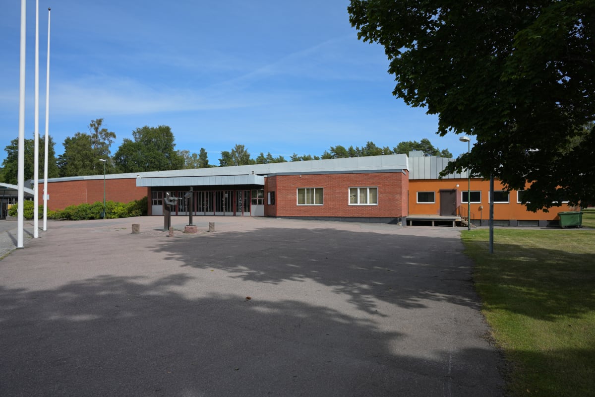 En stor, röd tegelbyggnad med detaljer i orange och grått under en blå himmel. Framför byggnaden finns en asfalterad yta med tre vita flaggstänger, två skulpturer och buskage. Träd är synliga i bakgrunden och i bildens övre högra hörn.