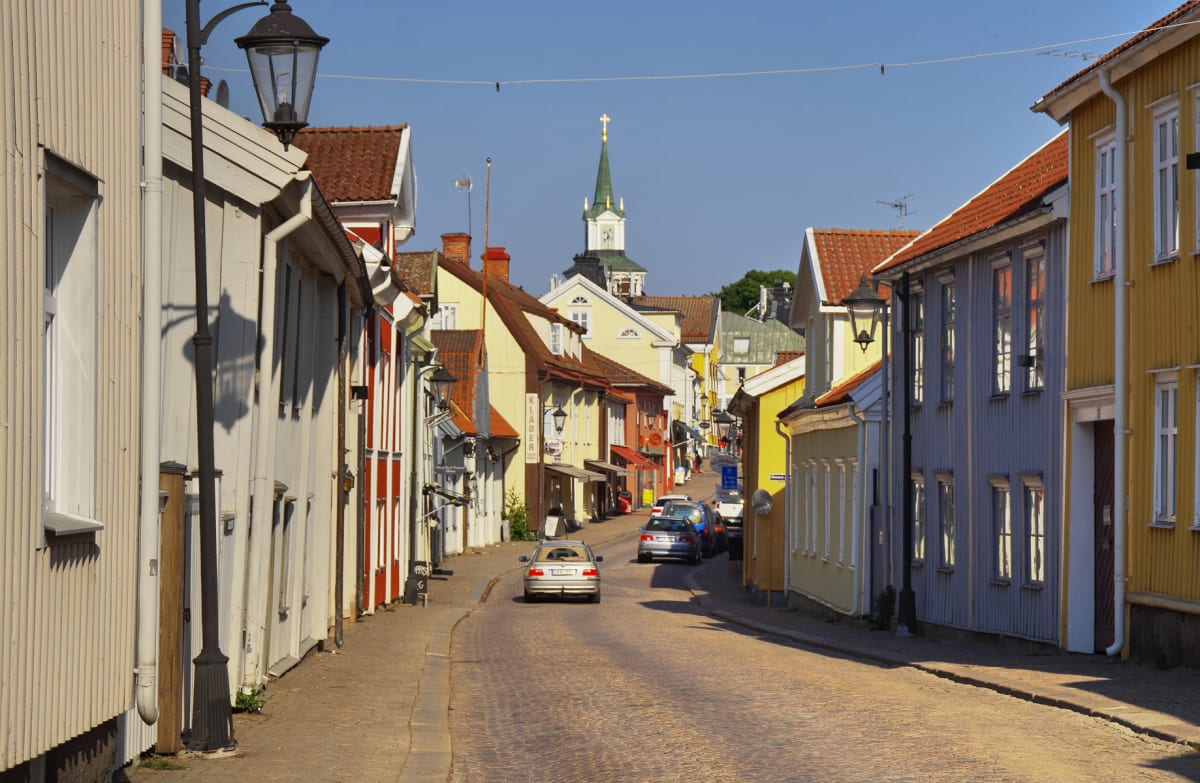 En gatsvy av en kullerstensgata kantad av flertalet äldre byggnader i olika färger, främst gult, vitt och rött. En kyrkspirat med en klocka syns i fonden. Flera bilar är parkerade eller kör på gatan och skuggor faller från byggnaderna. Himlen är klar