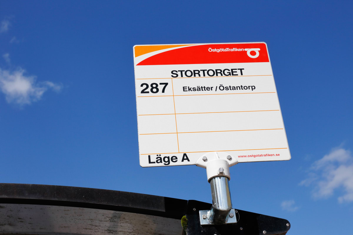 En gul och röd busshållplatsskylt från Östgötatrafiken, med texten "Stortorget", "287 Eksätter / Östantorp" och "Läge A", mot en klarblå himmel med några vita moln. Skylten är monterad på en metallstolpe och syns delvis ovanför en mörk struktur.