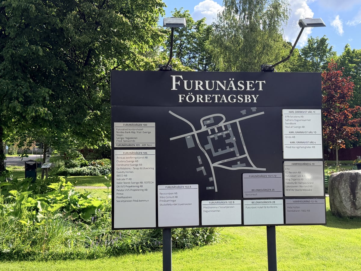 En svart skylt för "Furunäset Företagsby" med en karta och listor över företag. Bakom skylten syns träd med gröna löv och blå himmel med några moln. Vid sidan av skylten syns gräs och buskage i förgrunden.