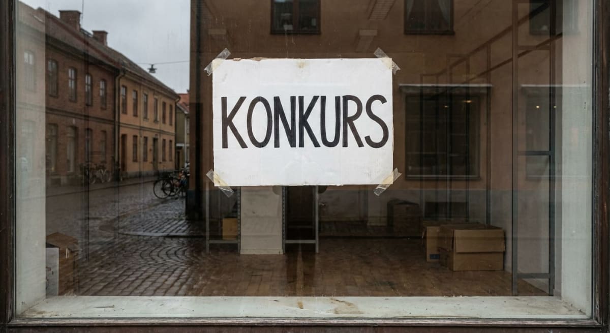 En vit skylt med texten "KONKURS" i stora, svarta bokstäver syns tejpad på ett skyltfönster. Genom fönstret reflekteras en kullerstensgata med byggnader och cyklar. Inne i butikslokalen syns tomma hyllor och kartonger.