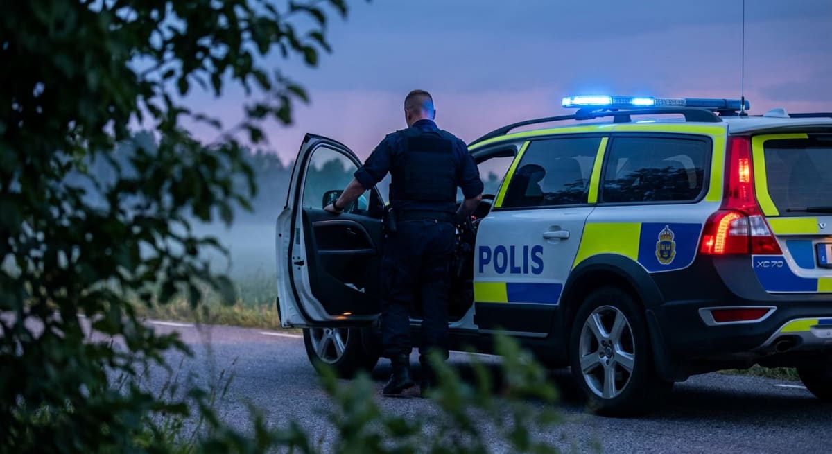En polisbil står med öppen bakdörr på en väg; blåljusen är tända och en person i polisuniform kliver ut. Runtomkring syns grönska och en gräsbevuxen dunge, vilket indikerar att det är utomhus. Ljuset på himlen antyder att det kan vara skymning eller