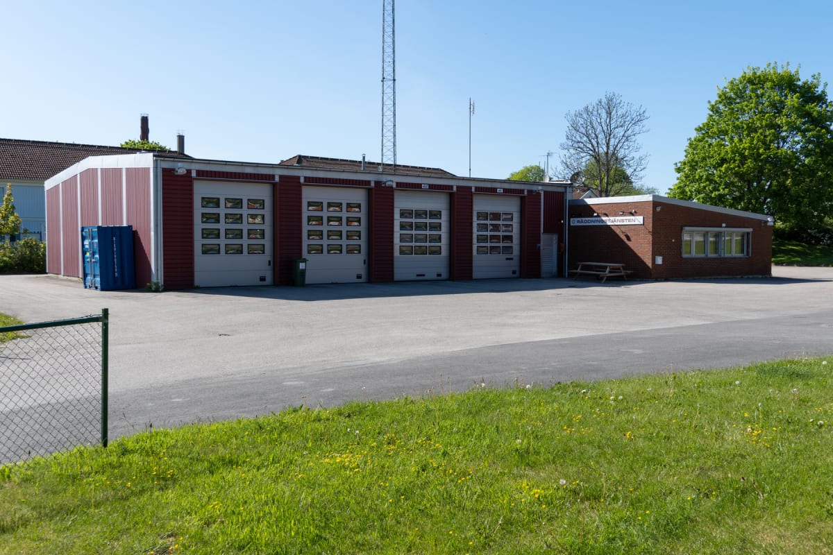 En röd tegelbyggnad med flera garageportar och ett fristående utrymme med en blå container utanför. En grön gräsmatta i förgrunden och träd samt en mast syns i bakgrunden. Ett picknickbord står vid entrén.