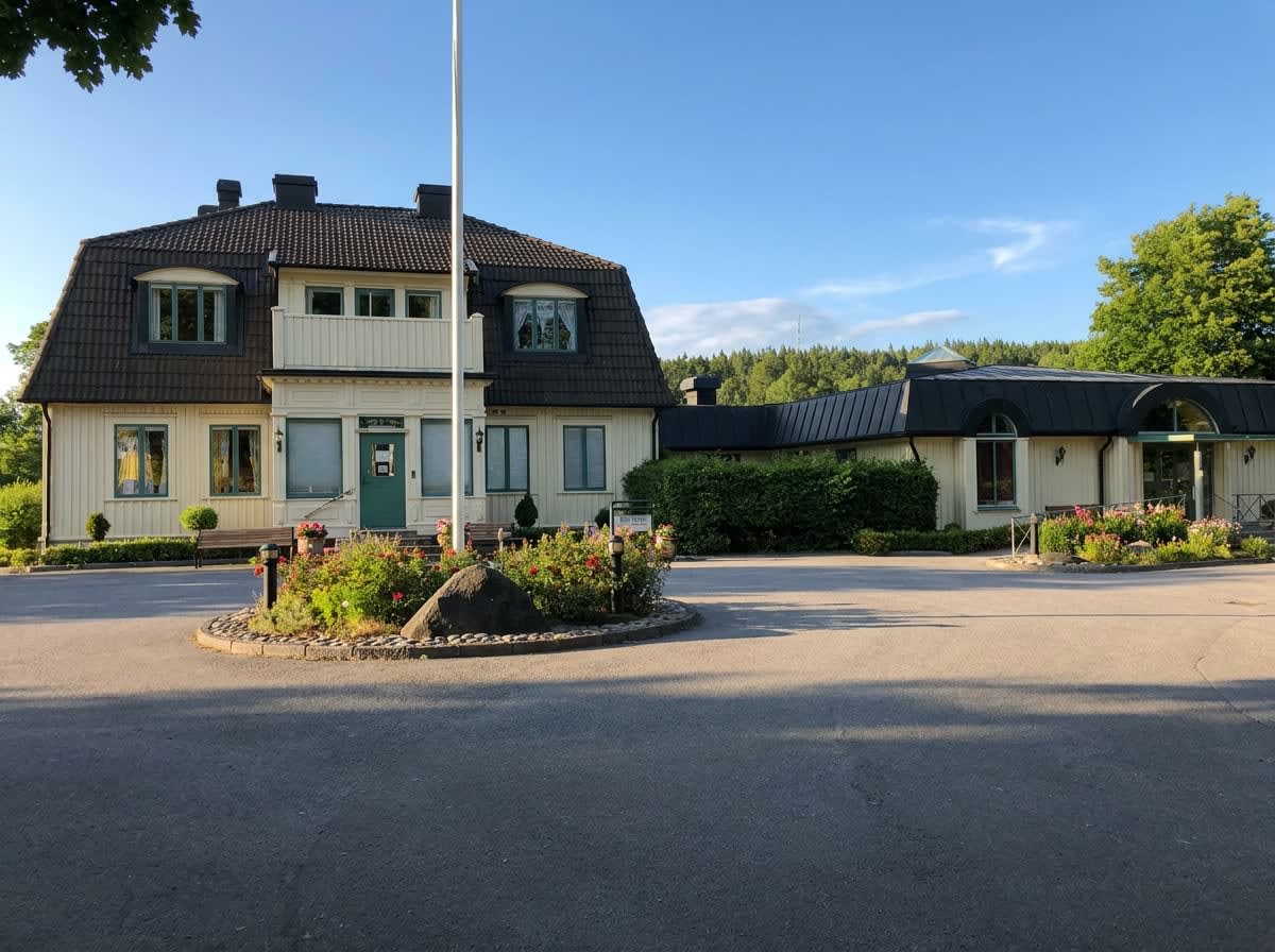 En stor, ljusgul byggnad med mörkt tak och flera fönster, omgiven av en asfalterad yta och planteringar med blommor. Framför byggnaden finns en flaggstång, buskar och träd. I bakgrunden syns skog och en blå himmel med några moln.