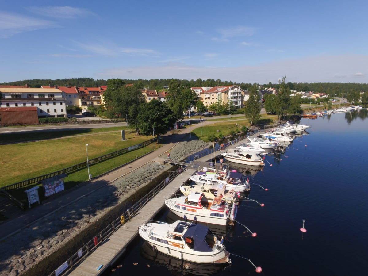 En högvinklad bild som visar en småbåtshamn med flera båtar förtöjda vid bryggor. I bakgrunden syns skogen och bebyggelse, inklusive flerbostadshus längs en gata med en trottoar och en gräsyta. Himmeln är blå med några lätta moln.