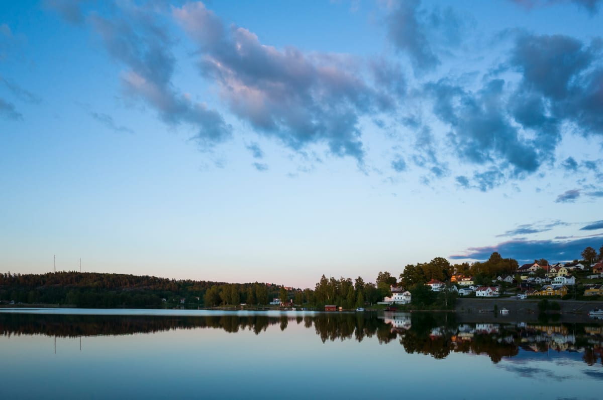 En vidvinkelbild tagen vid skymning, som visar ett lugnt vatten. På horisonten syns en skogsklädd kulle på vänster sida och en rad färgglada hus med röda tak på höger sida. Dessa byggnader speglas tydligt i vattnet under en ljusblå himmel med moln.