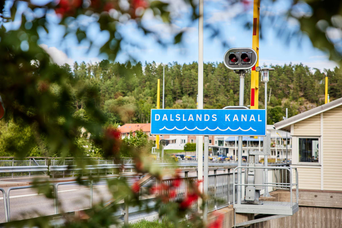 En bild tagen utomhus under dagtid, där en blå skylt med texten "DALSLANDS KANAL" i vit färg dominerar bilden. I bakgrunden syns skogklädda höjder, delar av en bro, samt byggnader och en sluss. Rödgröna lövverk i förgrunden ramar in bilden.