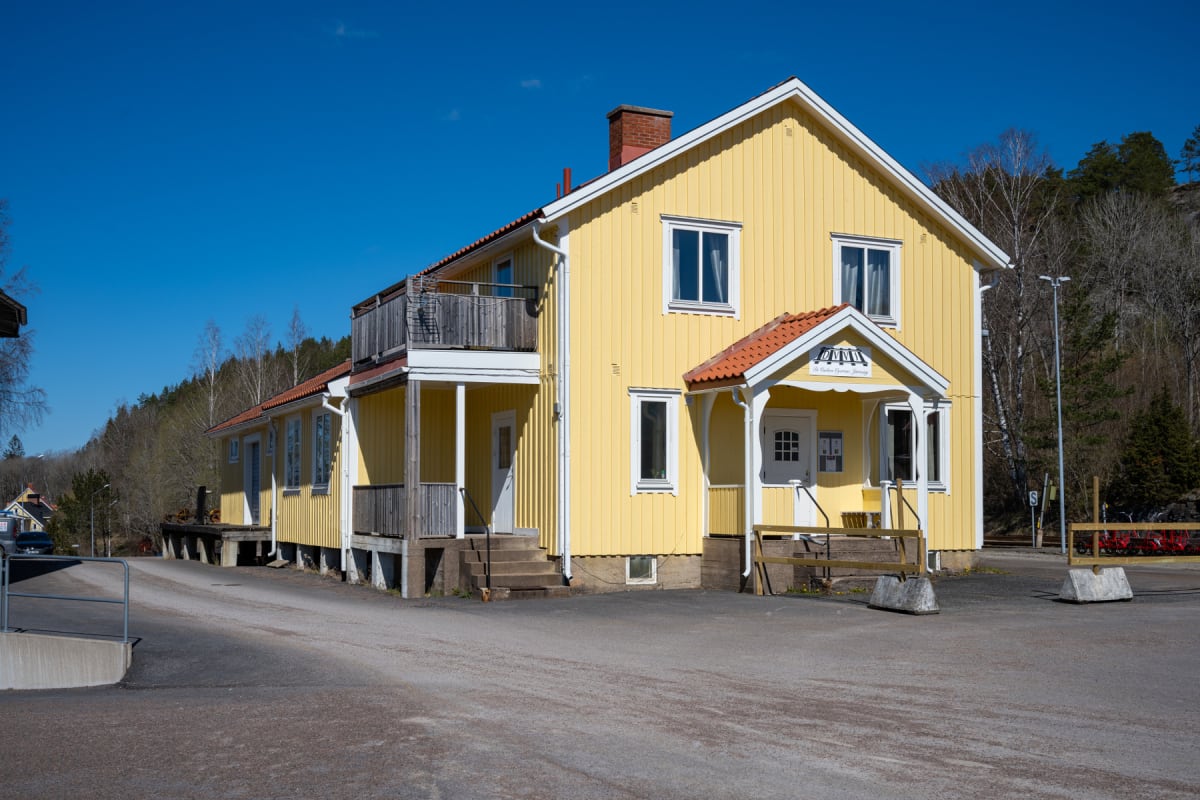 En gul byggnad med vita knutar och röda takpannor syns framifrån med en bred asfalterad yta framför. Byggnaden har flera fönster och dörrar samt en mindre balkong. Träd och skogsmark syns i bakgrunden under en klarblå himmel.