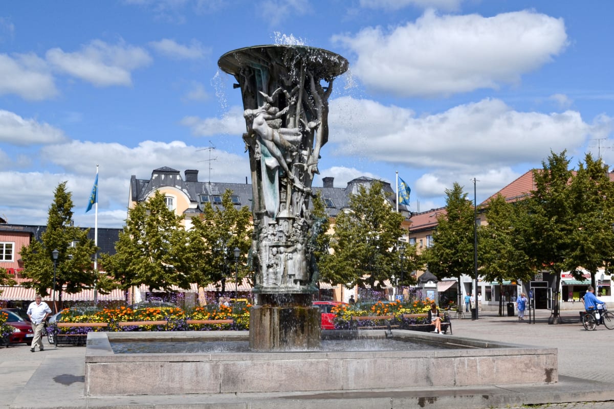 En stor bronsfontän står i mitten av bilden med vatten som sprutar ur toppen, omgiven av planteringar med färgglada blommor. I bakgrunden syns byggnader med olika tak och en ljusblå himmel med vita moln ovanför.