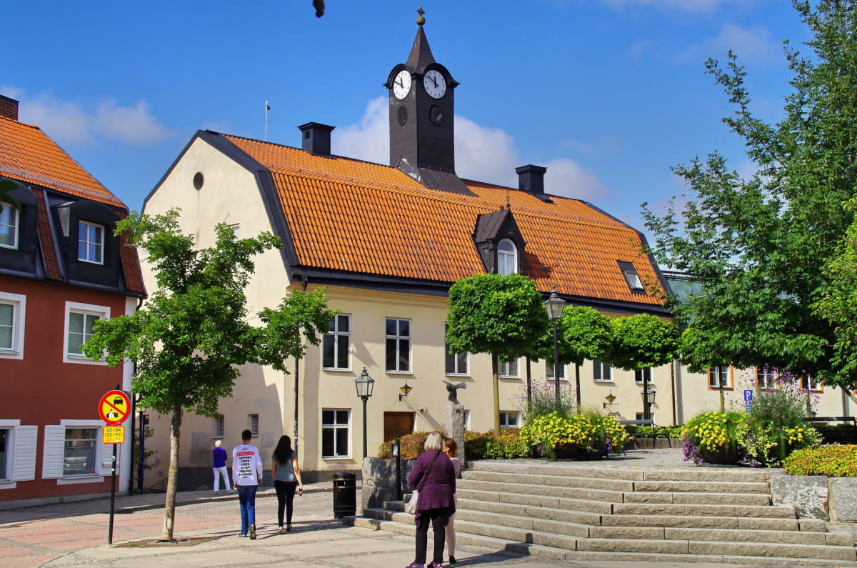 En bild en gata med flera personer, träd och byggnader med röda tak under en klarblå himmel. En av byggnaderna har ett klocktorn och sten trappor, blommor i planteringar och lyktstolpar syns.