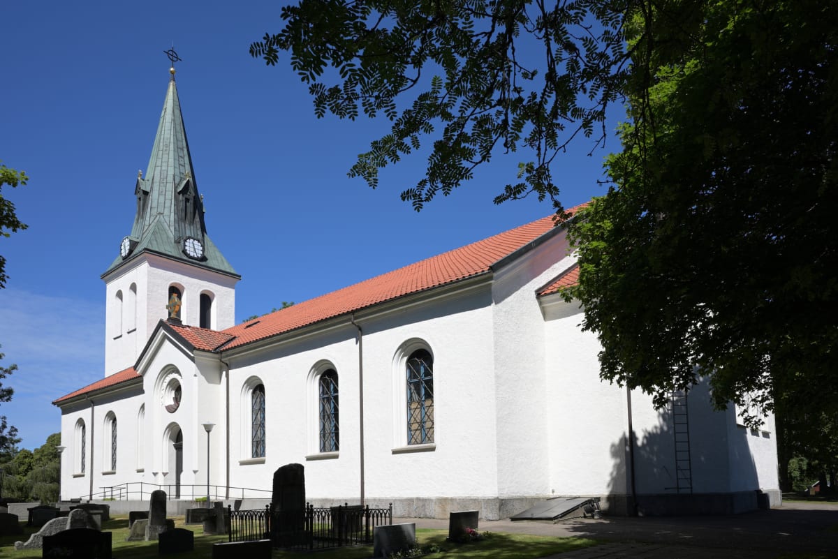 En vit kyrka med rött tegeltak och grön kyrkspira syns under en klarblå himmel. Framför kyrkan finns en kyrkogård med gravstenar och träd, varav ett kastar skugga över kyrkobyggnaden.