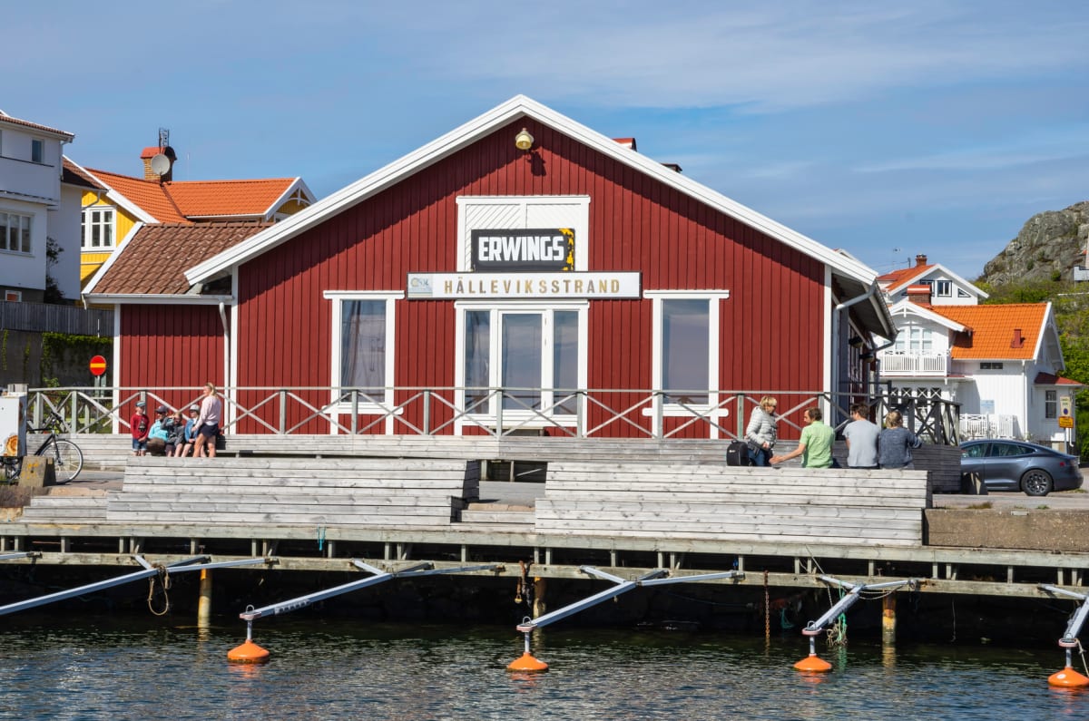 En röd byggnad med vita knutar och texten "ERWINGS HÄLLEVIKSSTRAND" på fasaden, belägen vid vattenbrynet. Flera personer syns sitta på en träbrygga framför byggnaden. I förgrunden syns vatten med orangea bojar och i bakgrunden bostadshus och en klipp