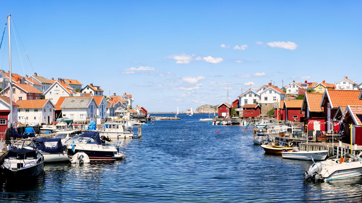 En vidvinkelbild som visar en smal vattenpassage med ett flertal båtar förtöjda vid bryggor på båda sidor. Hus i olika färger, främst vita och röda med tegeltak, kantar vattnet. I bakgrunden syns öppet hav och flera segelbåtar under en klarblå himmel