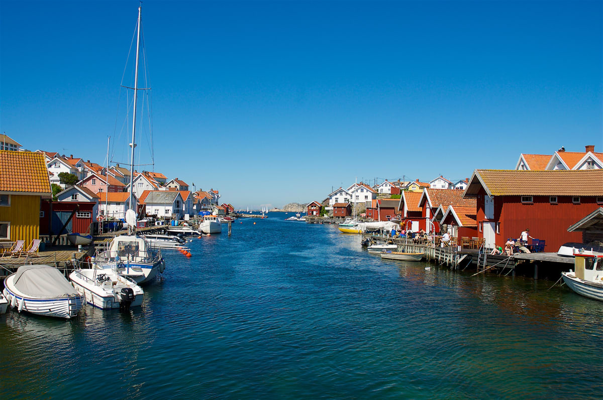 En bild över en blå vattenväg med byggnader och båtar på båda sidor. På vänster sida syns hus i olika färger, inklusive gult och rött, samt flera förtöjda båtar. På höger sida står röda sjöbodar med ljusa tak, där människor syns sitta på terrasser. I