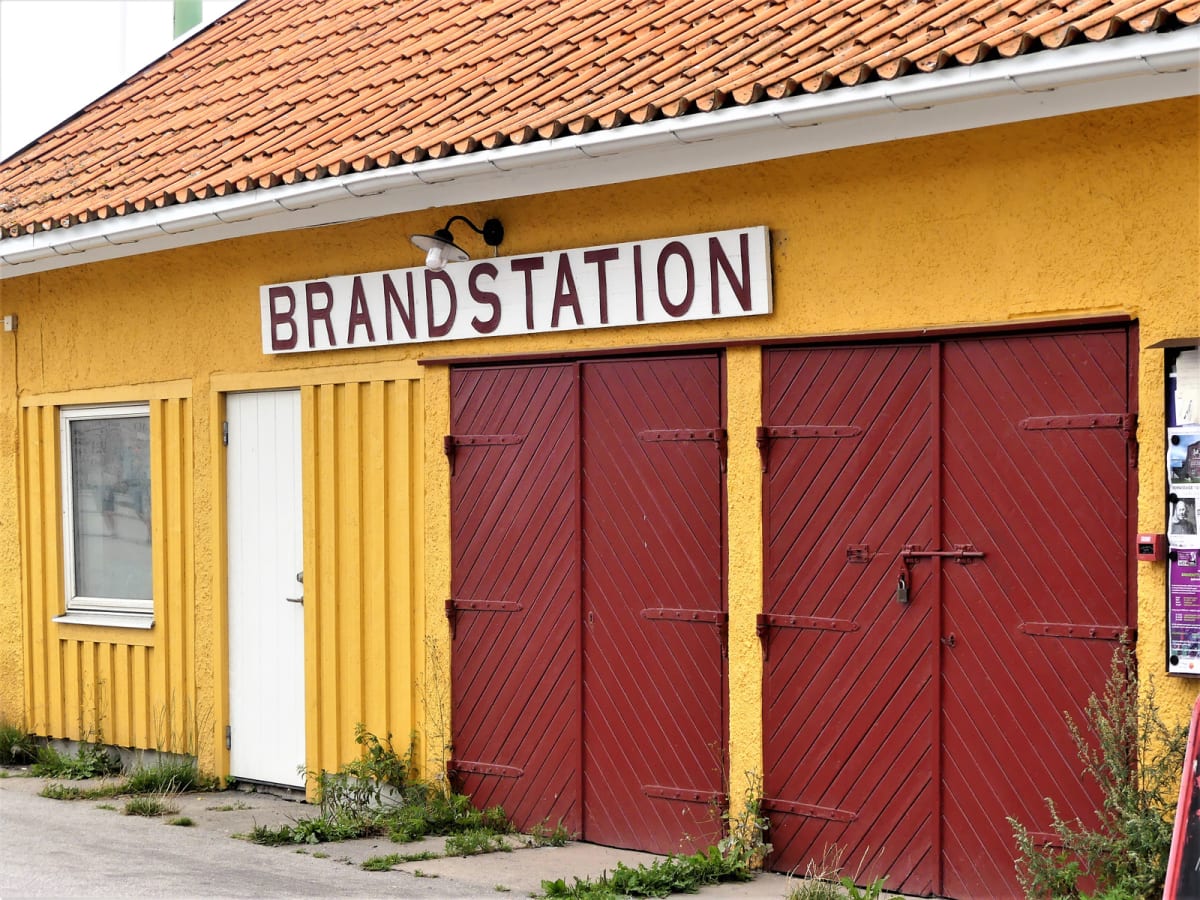 En gul byggnad med rött tak och röda dörrar syns. På fasaden finns en skylt med texten "BRANDSTATION". Till vänster finns ett fönster och en vit dörr. Två stora röda portar med diagonala ribbor är stängda. Ogräs växer vid byggnadens fot.