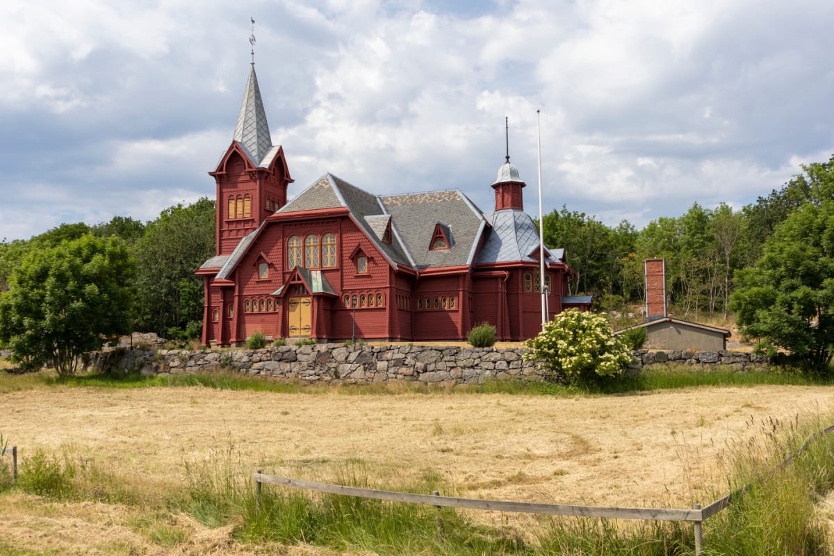 En röd träkyrka med grått tak och en spetsig spira står på en kulle. Framför kyrkan ligger ett torrt gräsfält med en låg trästaket i förgrunden. Grönskande träd syns runt kyrkan och i bakgrunden. En mur omger kyrkobyggnaden.