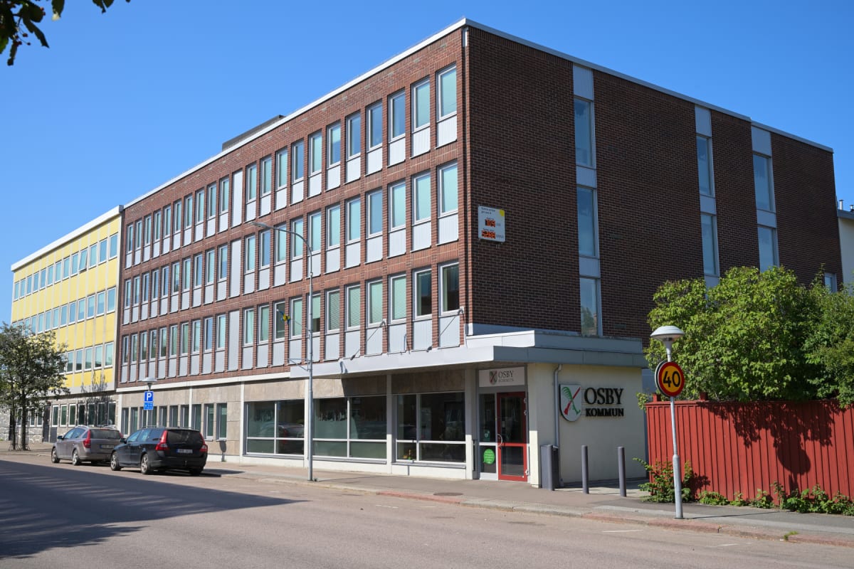 En stor, tegelröd byggnad syns från gatan under en klarblå himmel. Framför byggnaden finns parkerade bilar, gatubelysning och en hastighetsskylt som anger 40 km/h. Entrén till Osby kommun är också synlig på byggnadens fasad.