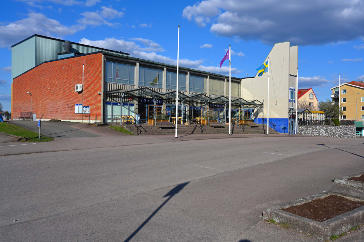 En modern byggnad med en fasad av tegel och en ljusblå korrugerad plåt på ena sidan. Flera ingångar är täckta av glastak, och det finns tre flaggstänger, varav en med svensk flagga och en med lila flagga. Framför byggnaden finns en asfalterad öppen y