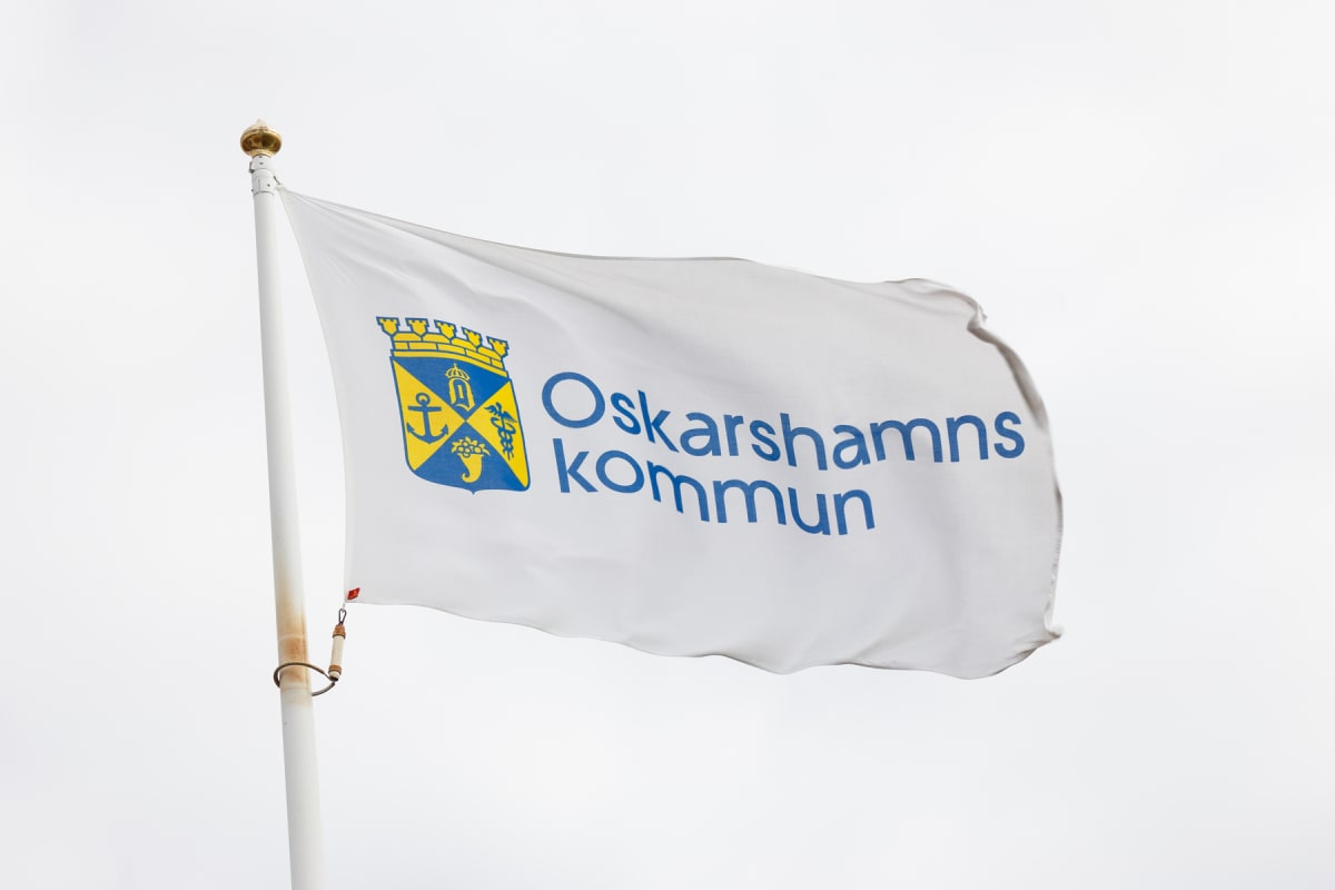 En vit flagga med blå och gul logotyp rör sig lätt i flaggstången under en ljus himmel. Logotypen innehåller texten "Oskarshamns kommun" i blått, samt ett stiliserat vapen i blått och gult med en krona, ett ankare och en vinge. Flaggstången är vit me