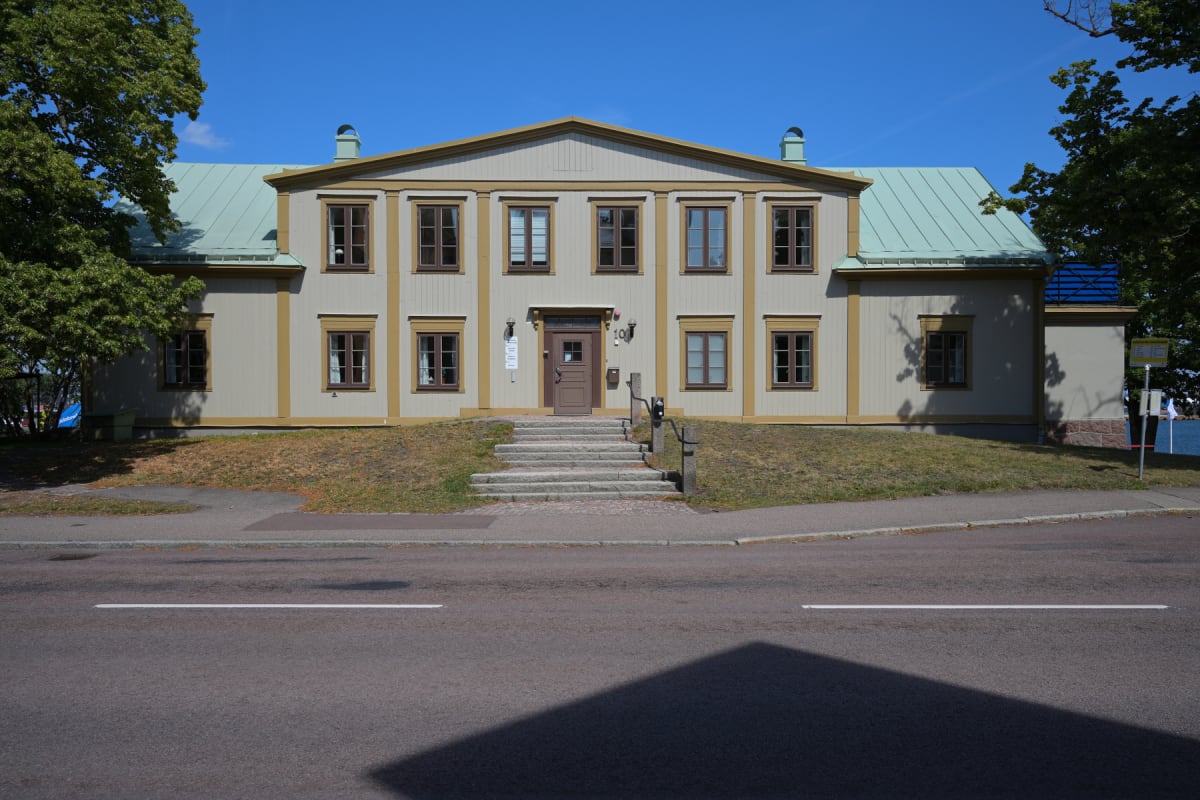 En stor, ljusbeige byggnad med mörkbruna fönster och dörr, samt ett grönt tak, står på en gräsklädd sluttning. Framför byggnaden finns en stentrappa som leder upp till entrén. Framför sluttningen löper en asfalterad väg med en vit linje. Skugga falle