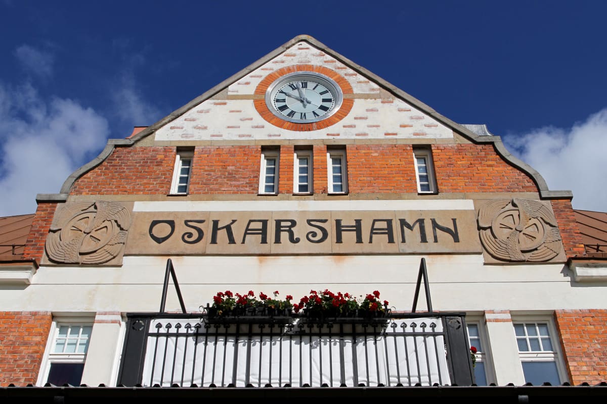 En fasad på en röd tegelbyggnad med vita detaljer och en stor urtavla högst upp i gaveln. Under urtavlan finns flera fönster och en skylt med texten "OSKARSHAMN". En balkong med svarta räcken och röda blommor syns i förgrunden.