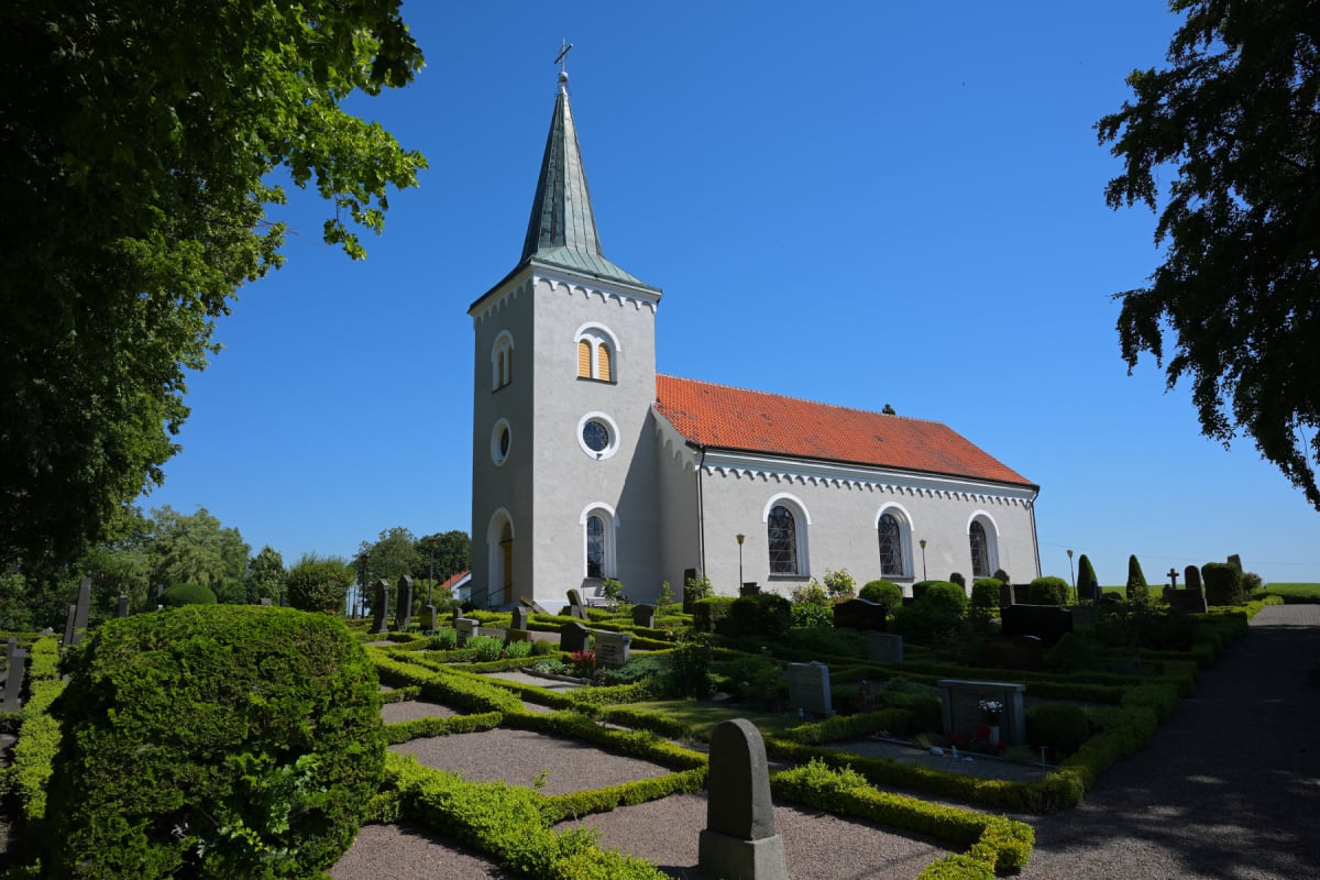 En kyrka belägen på en kyrkogård under en klarblå himmel. Kyrkan har ljusgrå puts, ett orange tegeltak och ett grönt torn med spira och kors. På kyrkogården syns gravstenar omgivna av formklippta buskar och grusgångar. Träd med gröna löv syns i bilde
