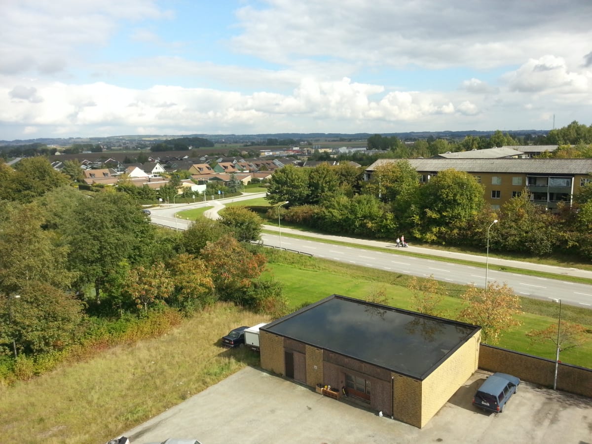 En högt tagen bild visar ett bostadsområde med hus och lägenhetskomplex under en delvis molnig himmel. En väg slingrar sig genom landskapet med gång- och cykelbanor. Två personer promenerar längs vägen. I förgrunden syns en tegelbyggnad med svart tak