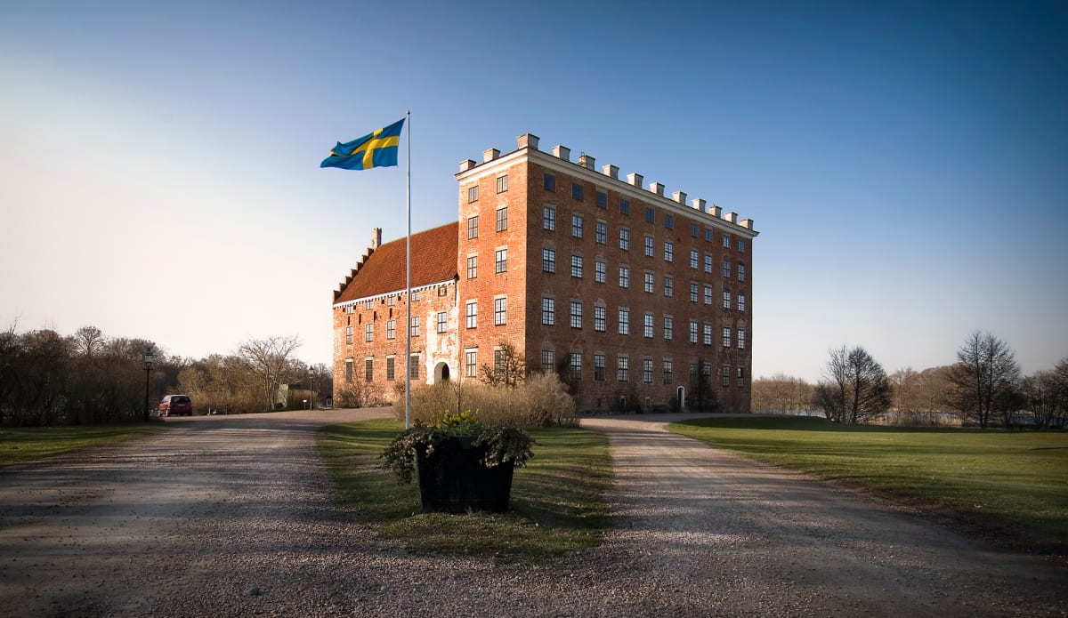 En stor, röd tegelbyggnad med många fönster står under en klarblå himmel. Framför byggnaden finns en svensk flagga på en flaggstång. En grusväg delar sig i förgrunden och gräsmattor breder ut sig jämte vägen. En röd bil är parkerad i bakgrunden till