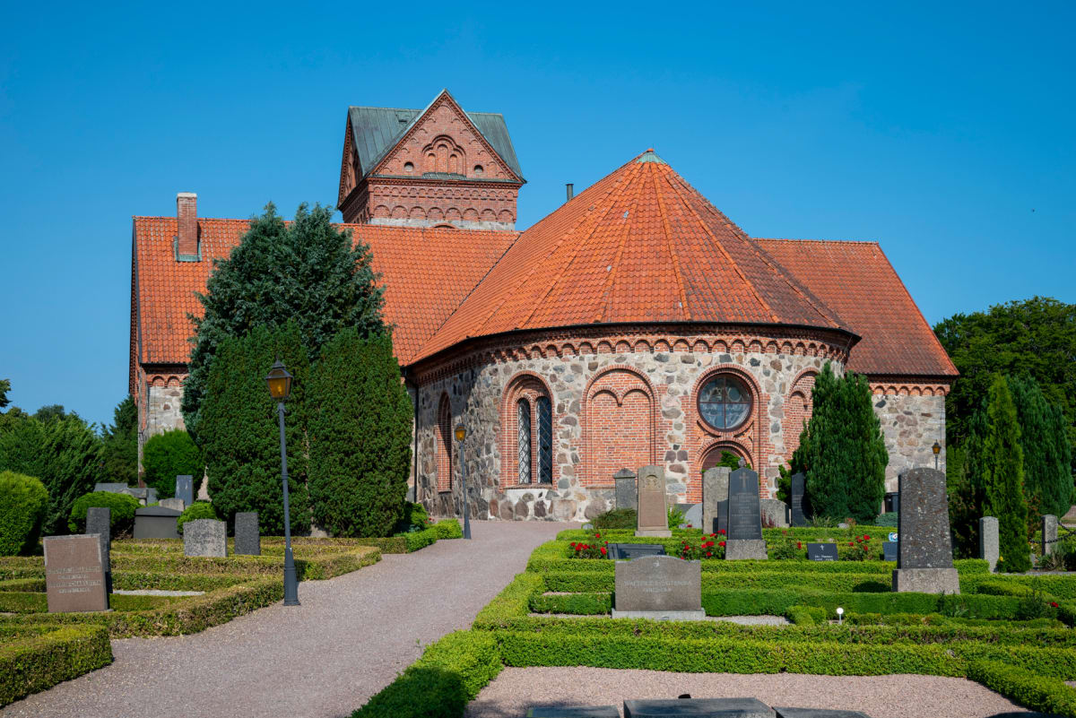 En kyrka med tegel- och stenfasad samt rött tegeltak under en klarblå himmel. På kyrkogården syns grusgångar, klippta häckar, träd, buskar, gravstenar och en lyktstolpe.