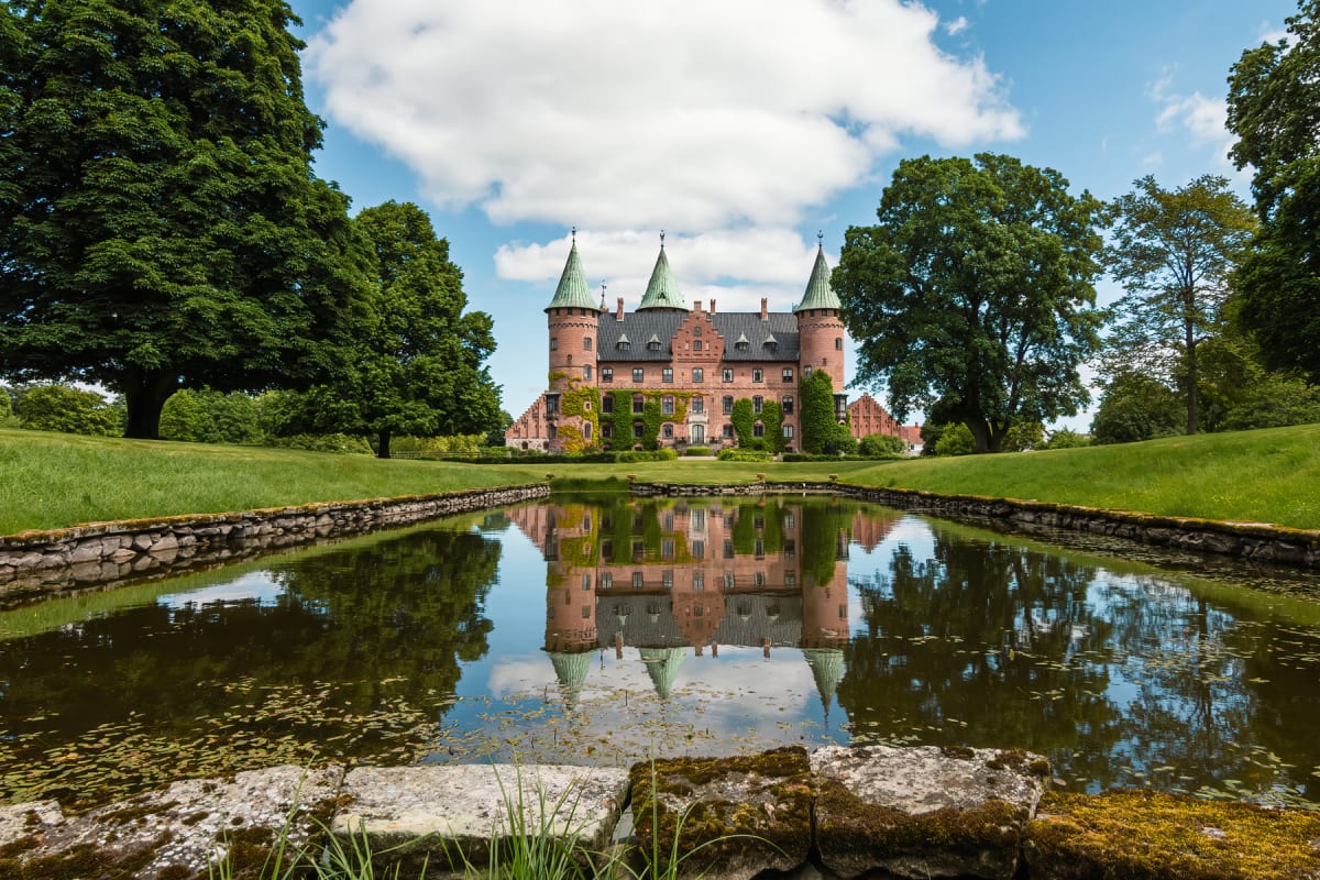 En stor, röd tegelbyggnad med flera torn och gröna spiror reflekteras i ett vattenområde i förgrunden, omgärdad av grönskande gräsmattor och stora träd under en blå himmel med vita moln.