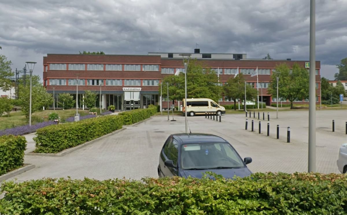 En bild som visar en tegelbyggnad med flera fönster under en molnig himmel. Framför byggnaden finns en parkeringsplats med flera parkerade bilar och planteringar med buskar och blommor. En gul skåpbil och en mörk personbil syns.